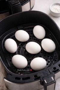 Seven whole eggs arranged in a single layer inside a black air fryer basket, ready to cook, with the perforated tray visible underneath.