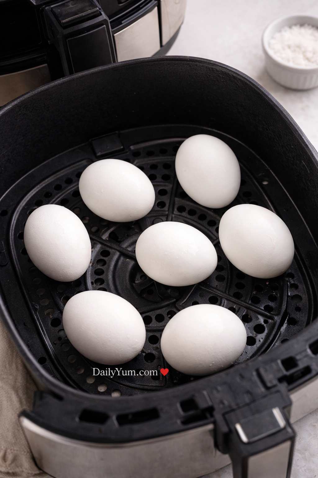 Seven whole eggs arranged in a single layer inside a black air fryer basket, ready to cook, with the perforated tray visible underneath.